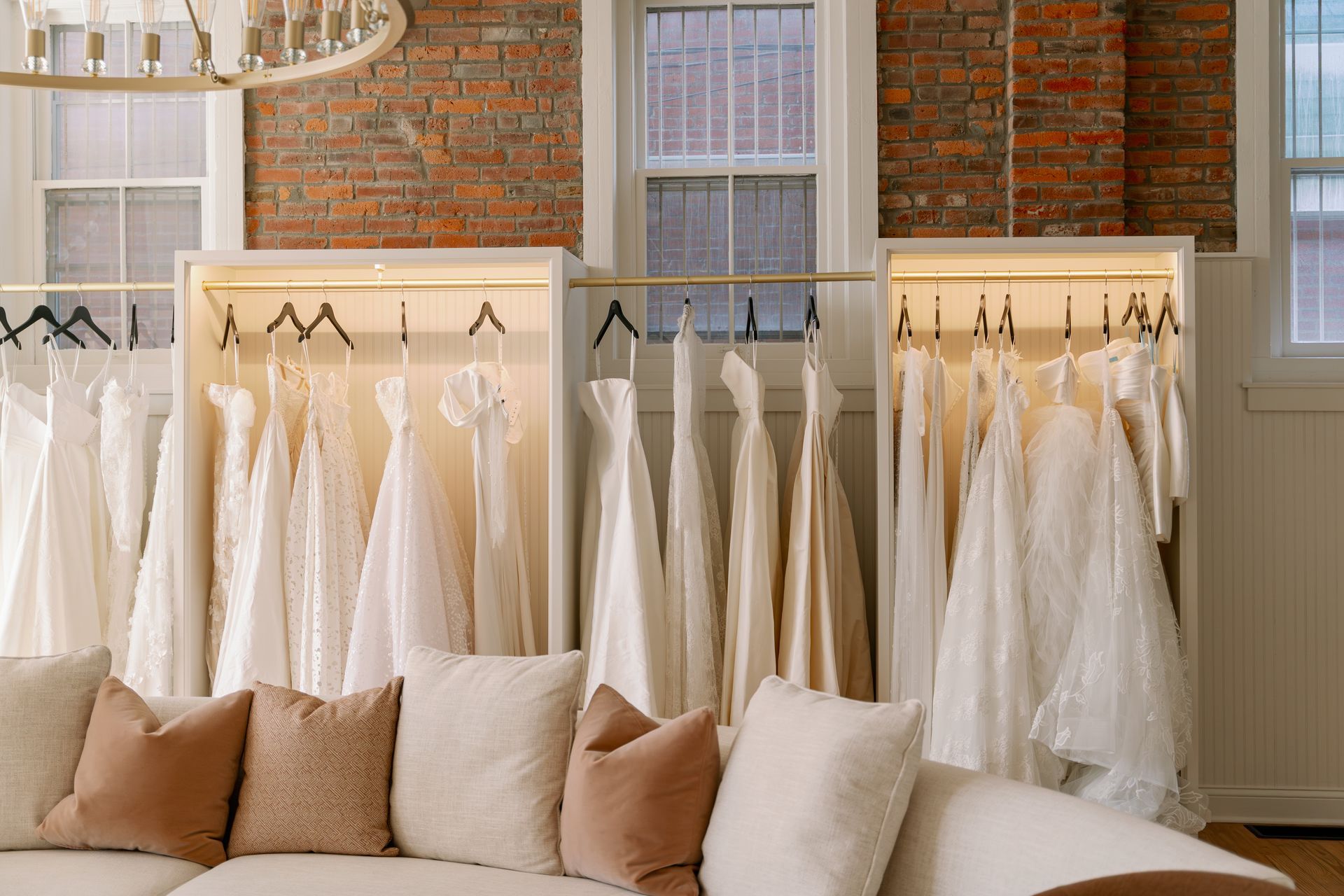 Wedding dresses displayed in a boutique. White gowns hang in illuminated cabinets above a white sofa with neutral pillows, against a brick wall.