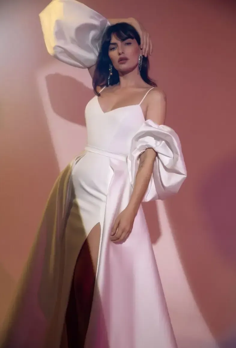 Woman in white wedding dress with puffy sleeves and thigh-high slit, posing in a studio.
