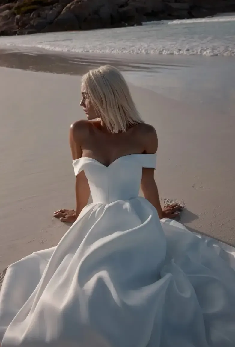 Woman in a white off-the-shoulder gown sits on a sandy beach, looking over her shoulder.