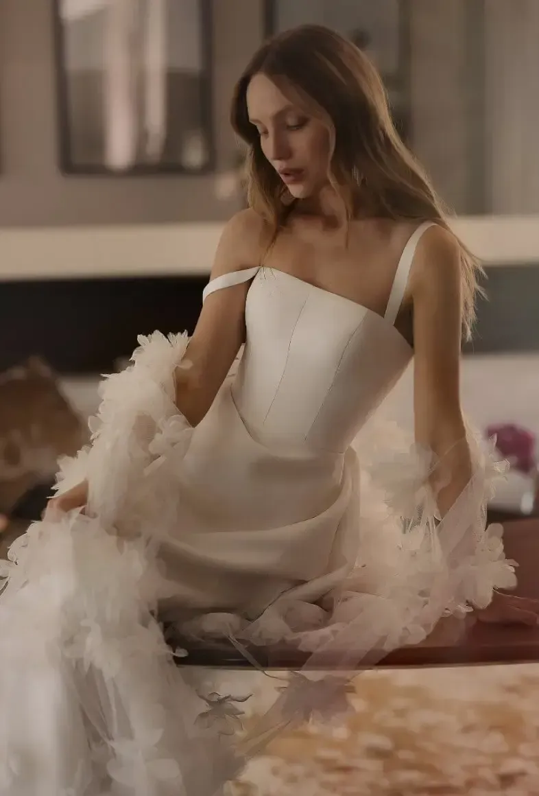 Woman in white wedding dress with ruffled train, leaning on a table, soft lighting.
