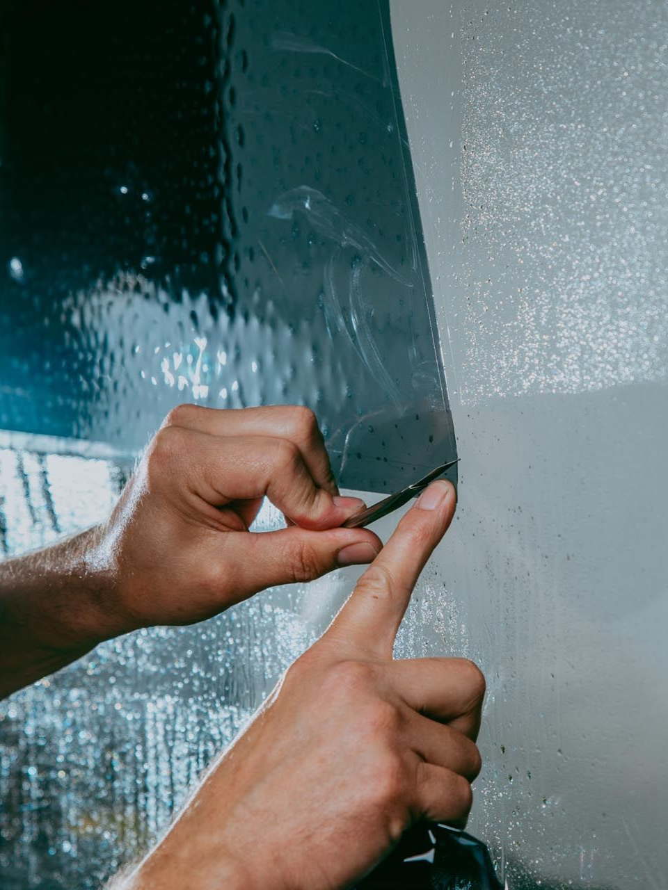 Hands using a knife to trim window tint film on a window, in a dim setting.
