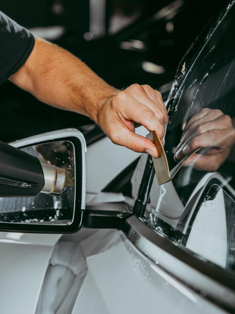 Person applying tint to a car window with a tool, white car, in a workshop.