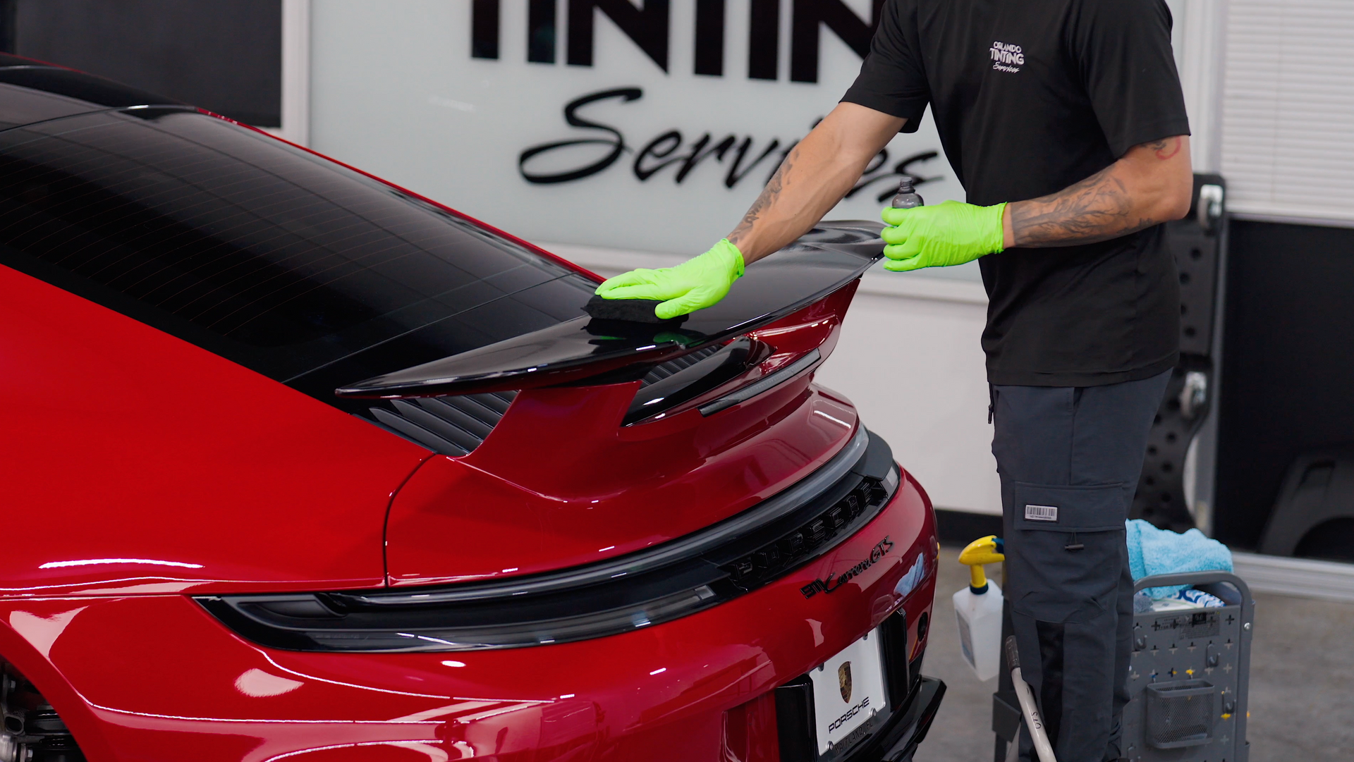 A person in green gloves cleans the spoiler of a red Porsche with a black rear wing.