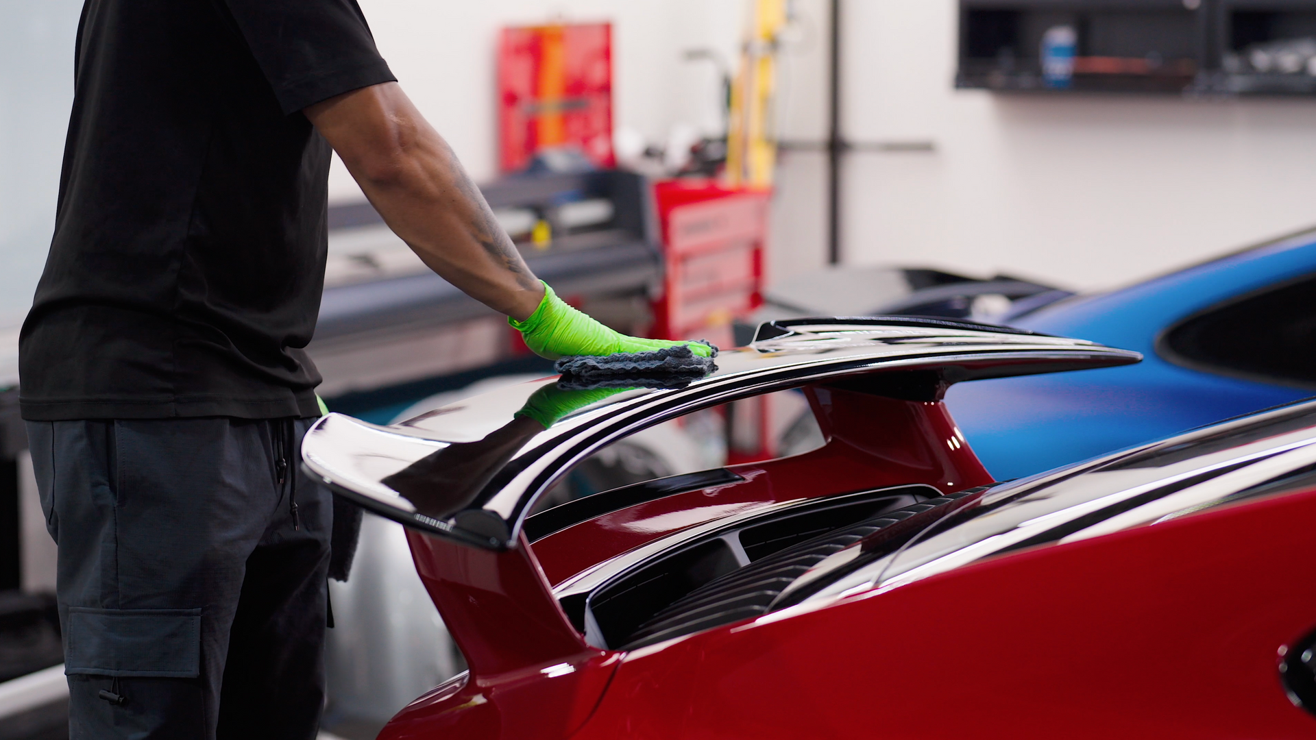 Person wearing a green glove cleaning a red car spoiler in a garage.