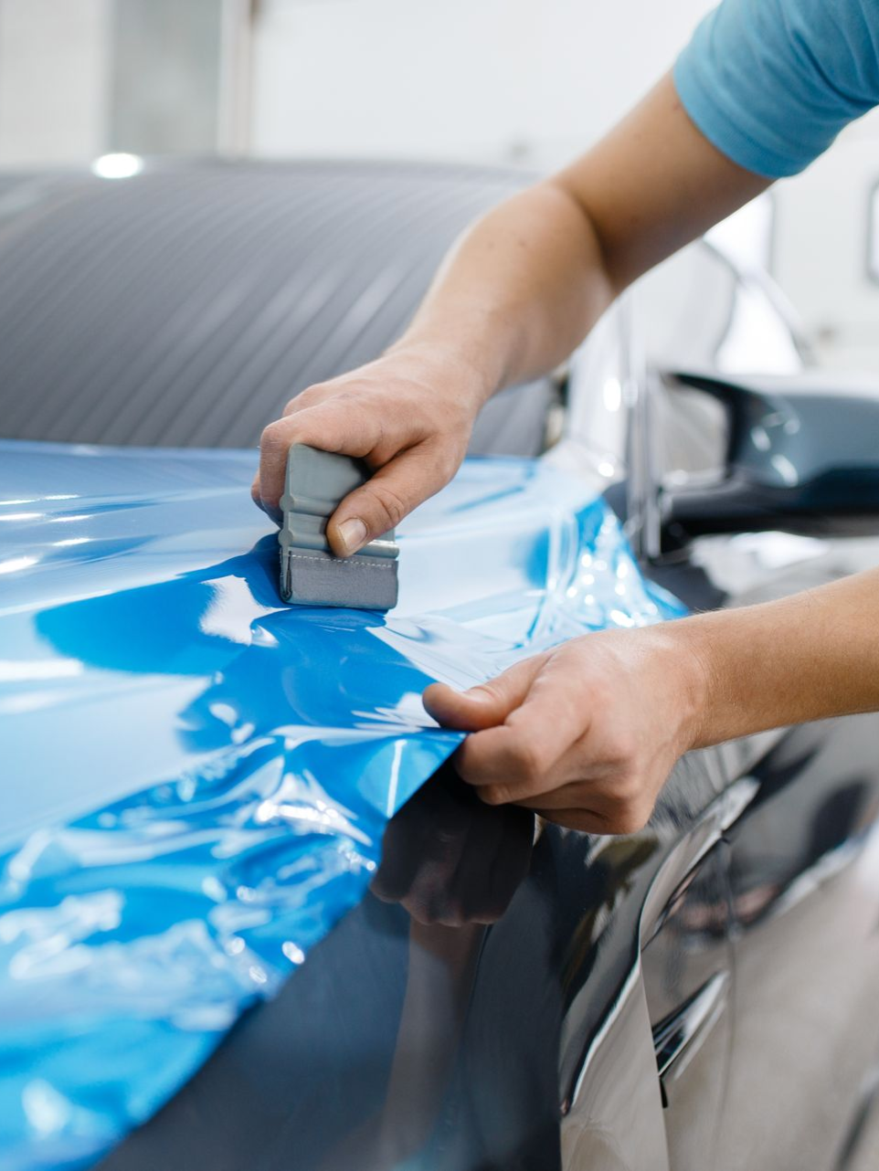 A person wrapping a blue film on a car's hood with a squeegee.