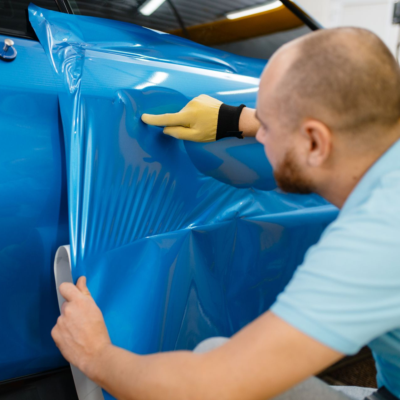 A man in a glove applying blue vinyl wrap to a car door, indoors.