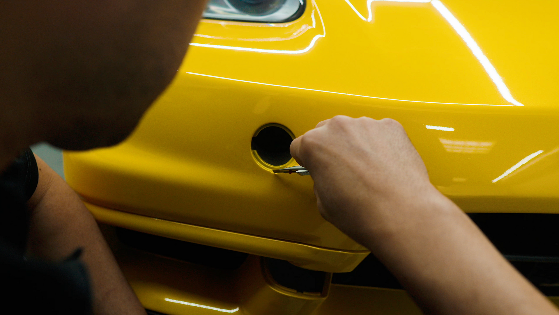 Person uses a tool to work on a yellow car's front bumper; close-up.