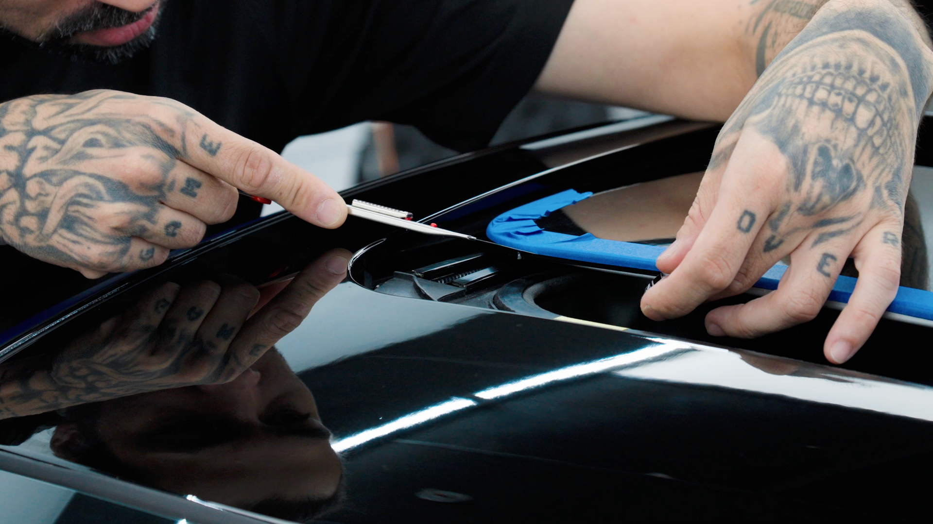 Person with tattoos trimming black car trim with a blade, using a blue guide.