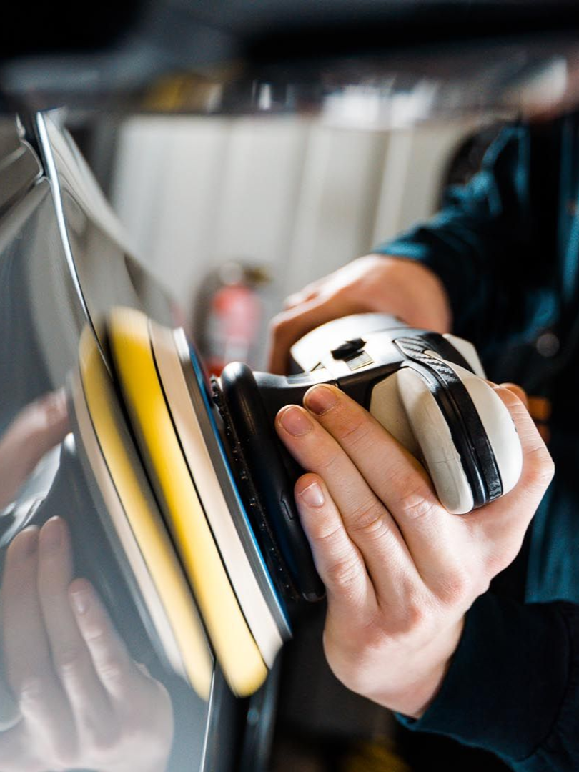 Person polishing car paint with an electric buffer, reflecting light, in a garage.