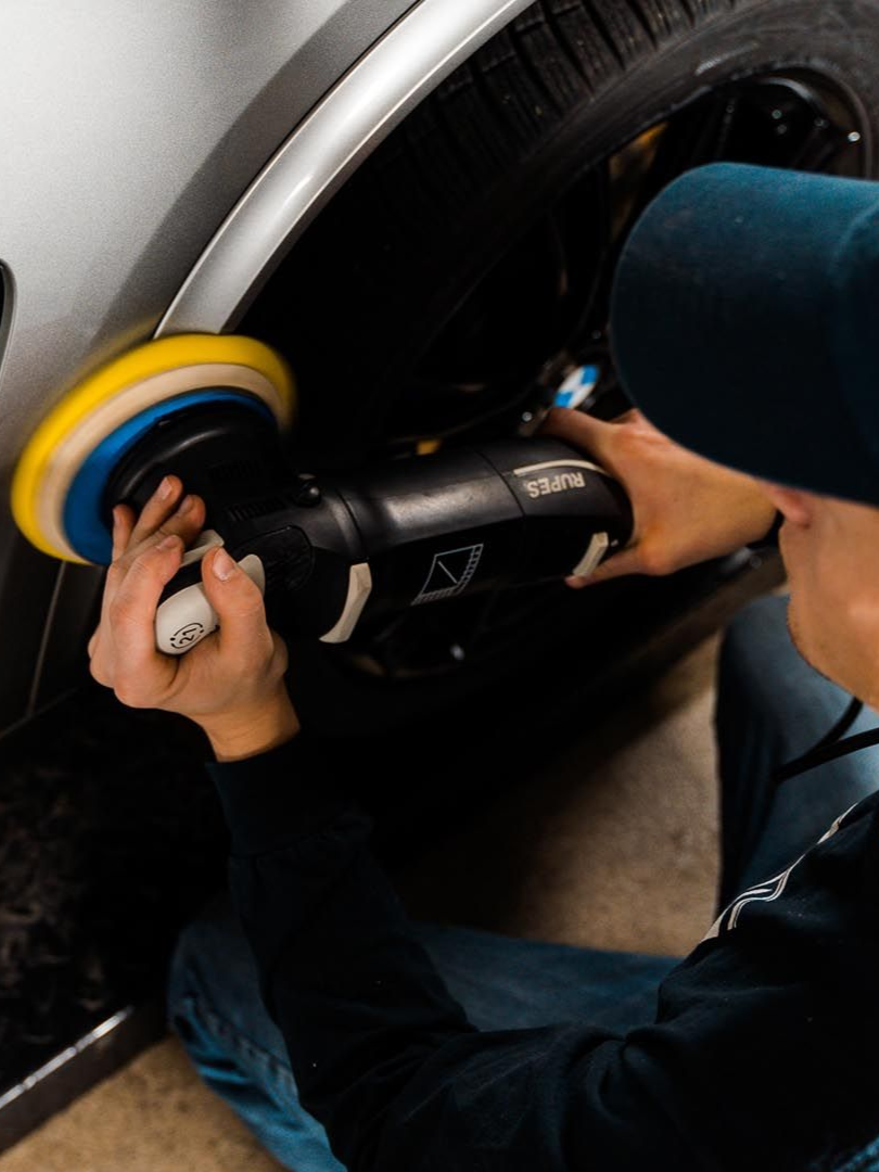 Person polishes a car with a power buffer, yellow and blue pad against silver body.