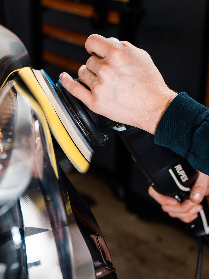 Person polishing a car panel with an electric buffer; black tool, yellow pad, shiny surface.