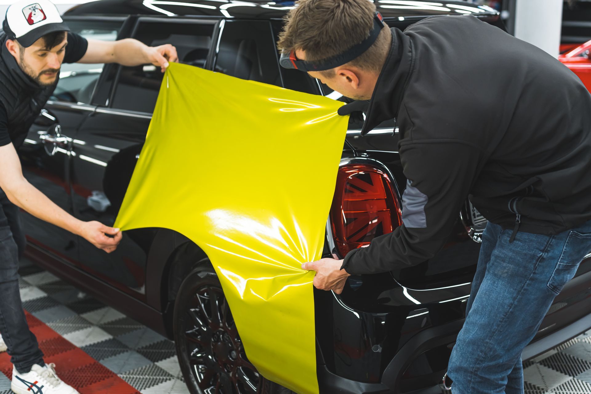 Two men installing yellow vinyl wrap on a black car, indoor setting.