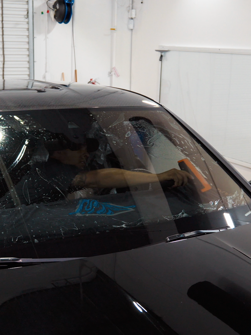 Car windshield being tinted; a person is smoothing film with an orange tool, indoors.
