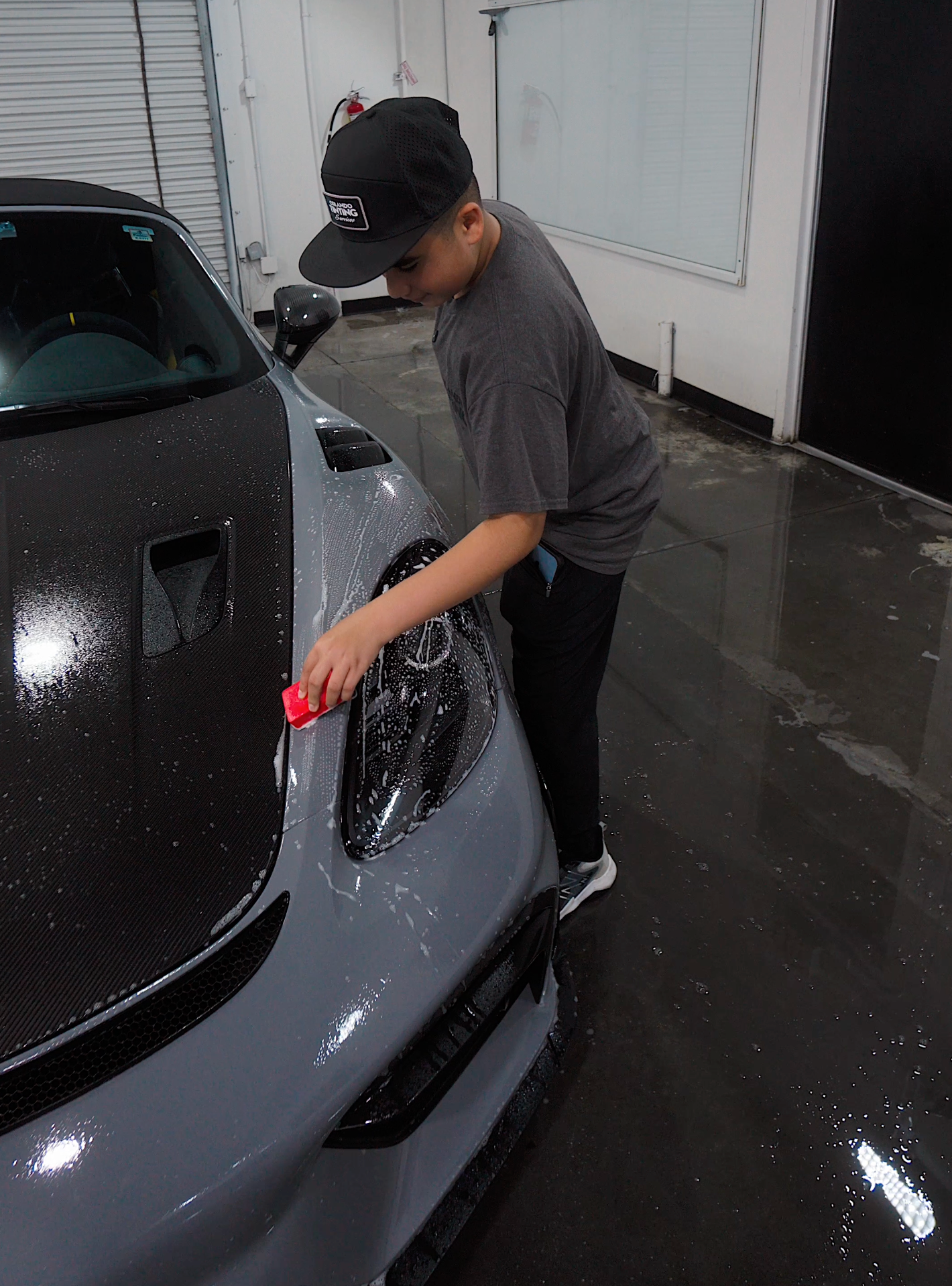 A person washes a gray car. They are wearing a hat and dark clothing in a well-lit space.