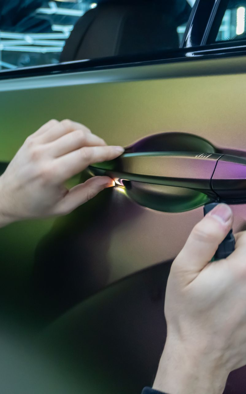 Hands wrapping iridescent vinyl around a car door handle on a vehicle.
