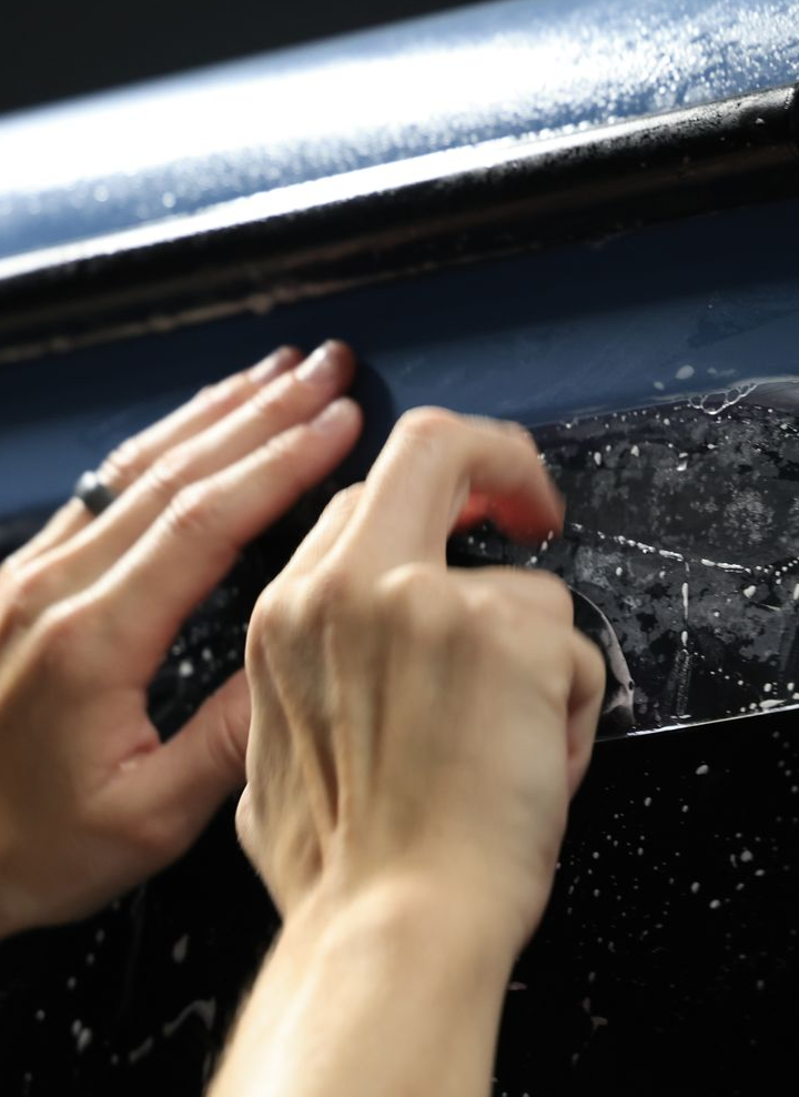 Hands applying window tint to a car's dark surface.