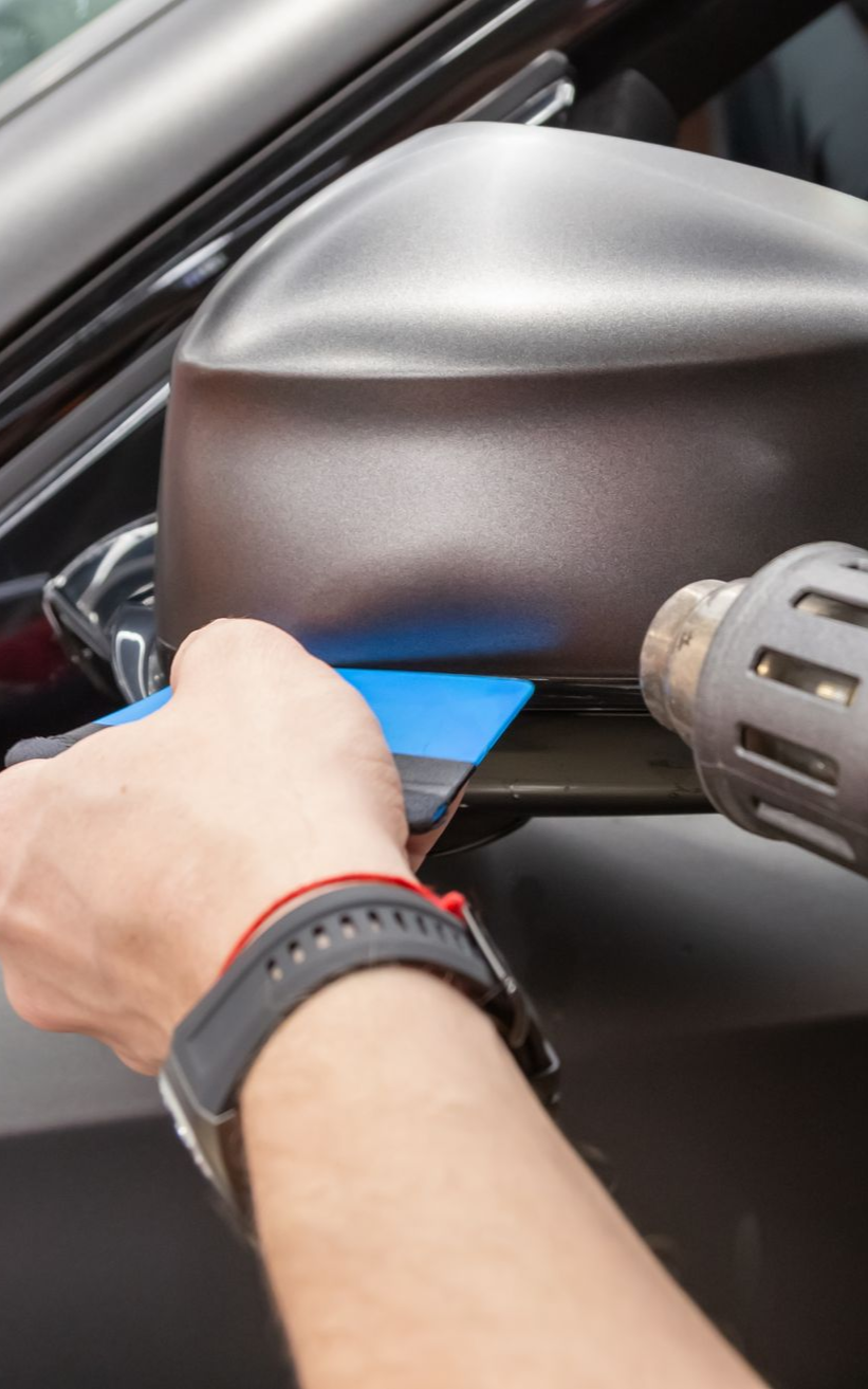 Person wrapping a car side mirror with brown vinyl, using a squeegee and heat gun.