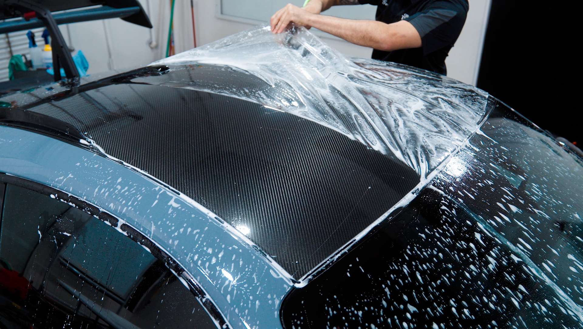 Person applying protective film to a car roof. Clear film over dark roof, soapy spray.