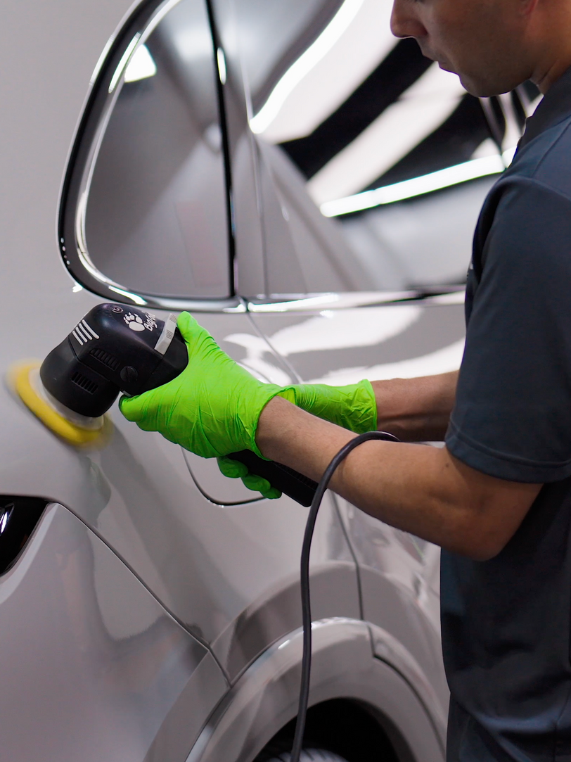 Man in green gloves polishing a white car with a black electric buffer.