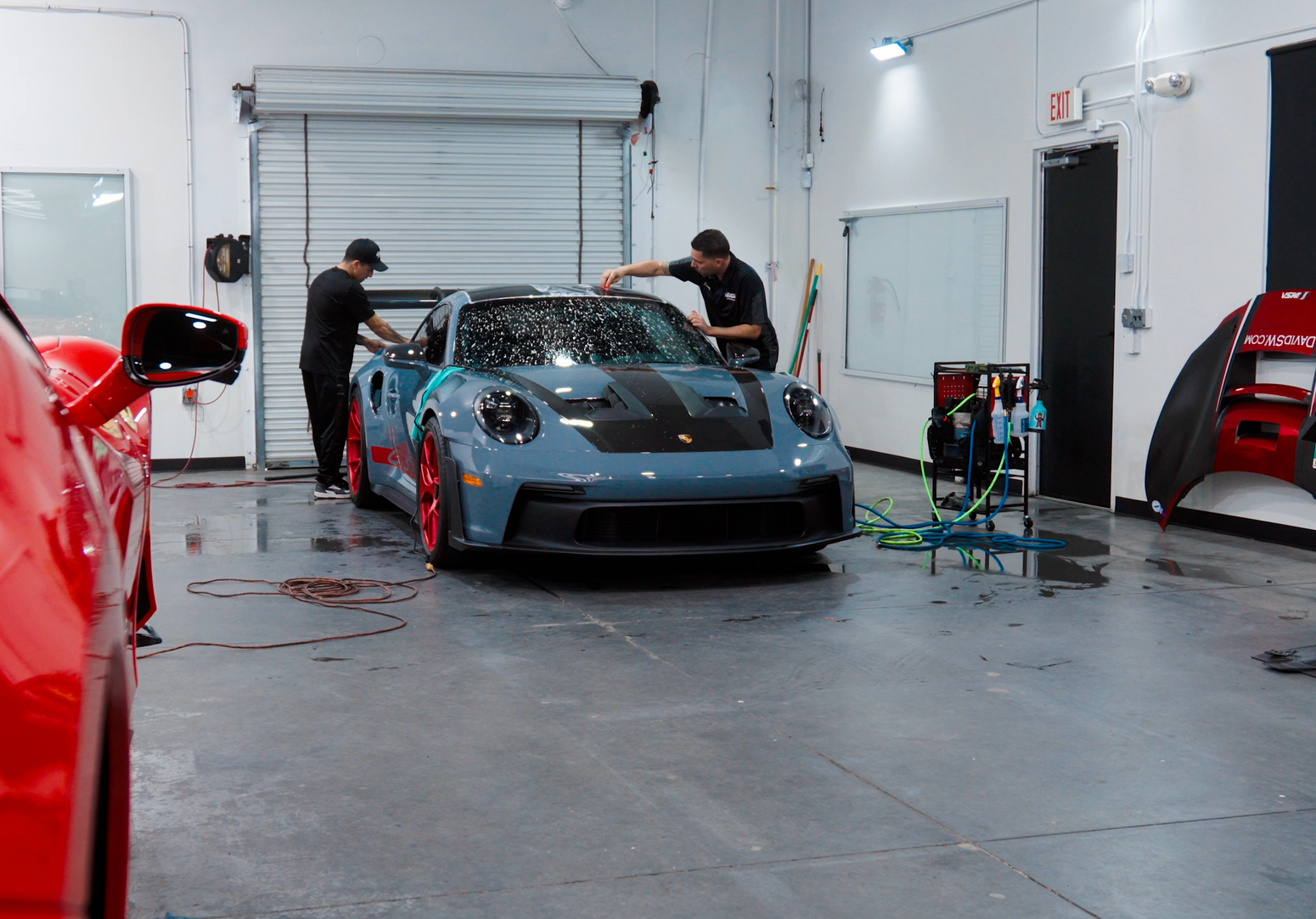 Two men washing a gray Porsche with red wheels inside a garage, a red car in the foreground.
