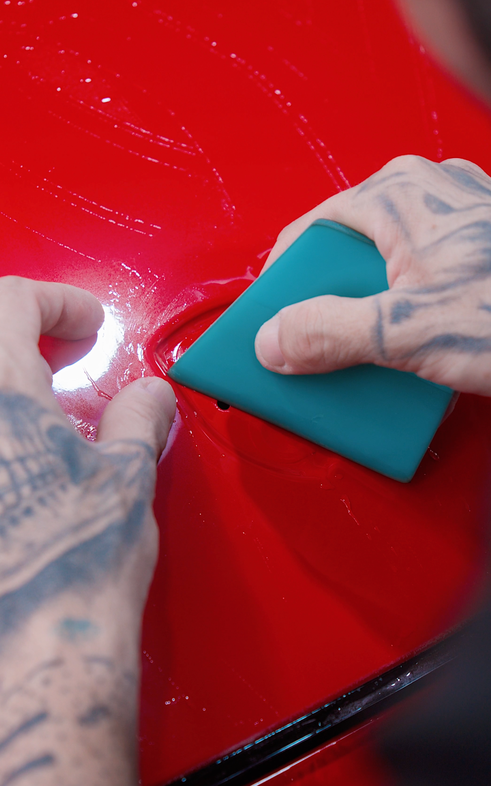Hands with tattoos smoothing film on red car with a teal squeegee.
