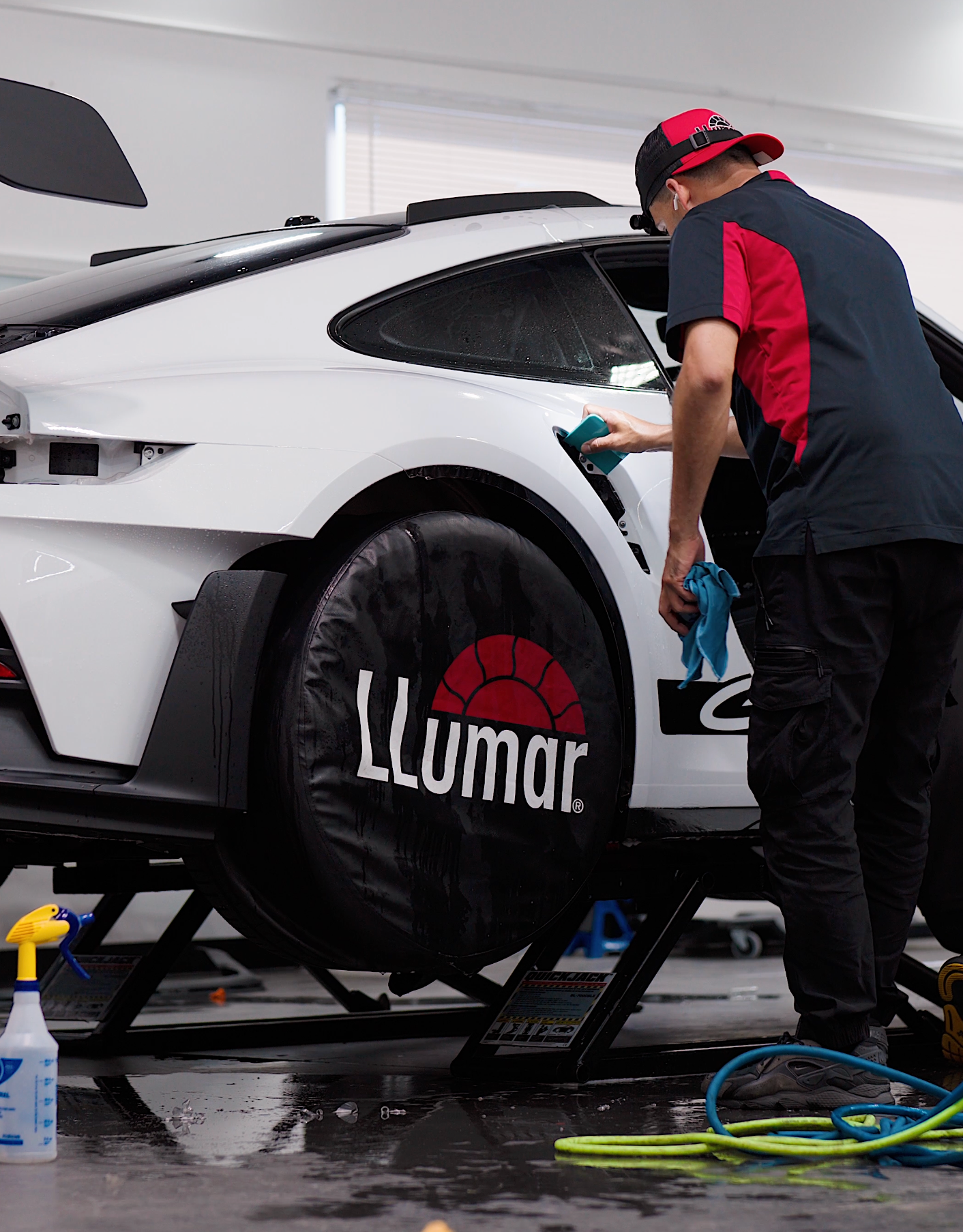 A person cleaning a white sports car's window. The car is on a lift with wheel covers labeled