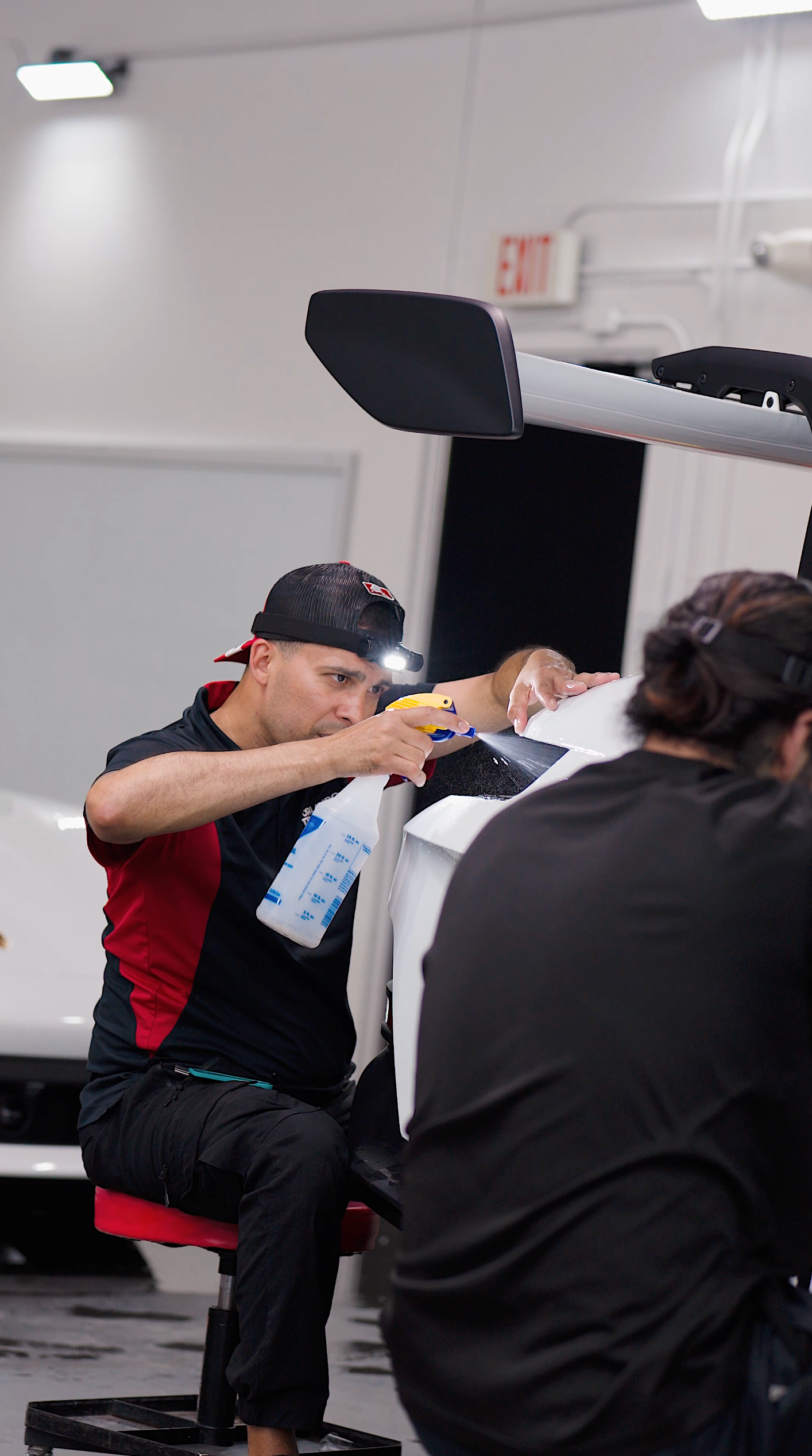 Man sprays a car spoiler, another person works nearby in a well-lit shop.