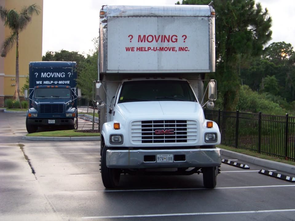 Two moving trucks parked in a parking lot. One white, one blue, both with moving company logo.