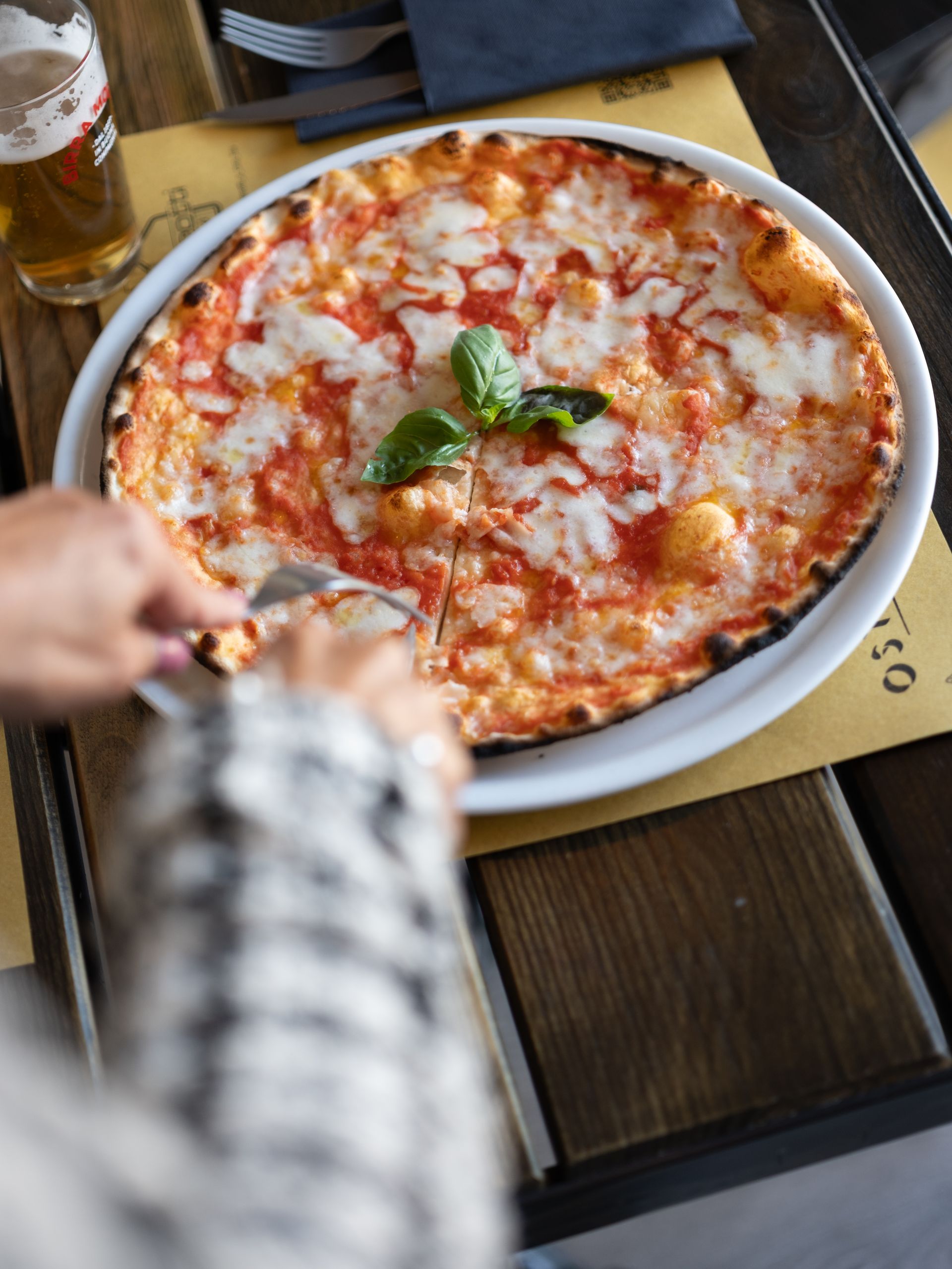 Persona che taglia una pizza Margherita con un coltello, guarnita con basilico, su un piatto e con una birra accanto.