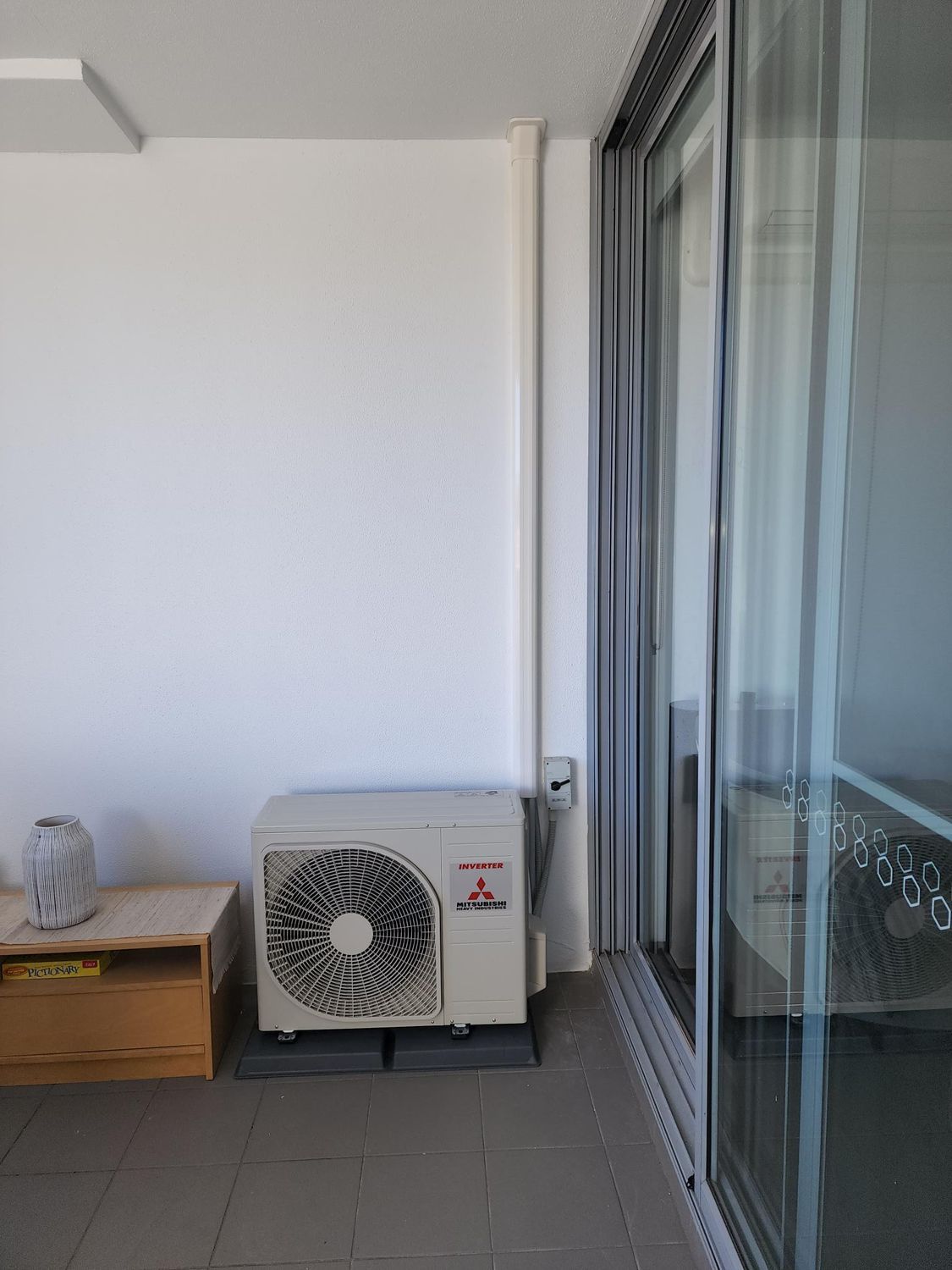 Air conditioning unit on a balcony next to glass doors, a wooden table, and a white wall.
