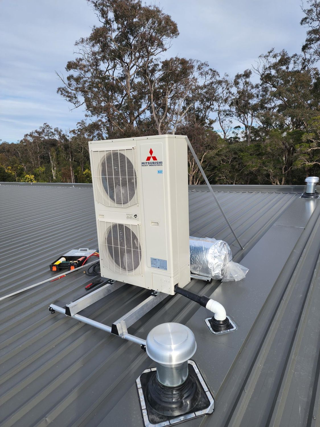 Mitsubishi air conditioning unit on a metal roof, with exhaust vent and background trees in Nowra