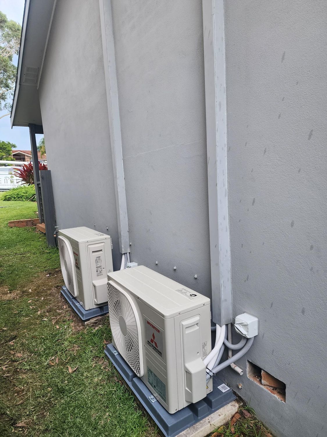 Two outdoor AC units on a gray wall. White tubes and electrical box visible. Green grass below.