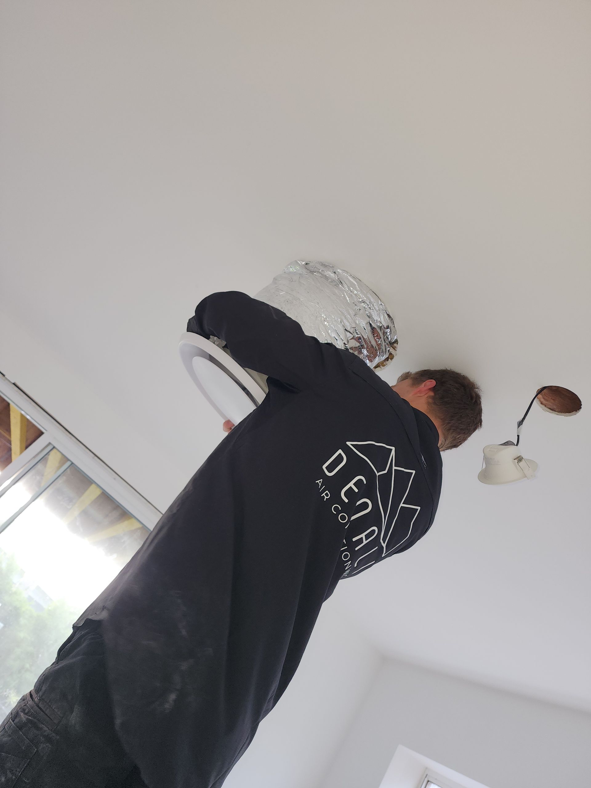 Man installing a ceiling light fixture. He is in a room with white walls, wearing a black shirt.