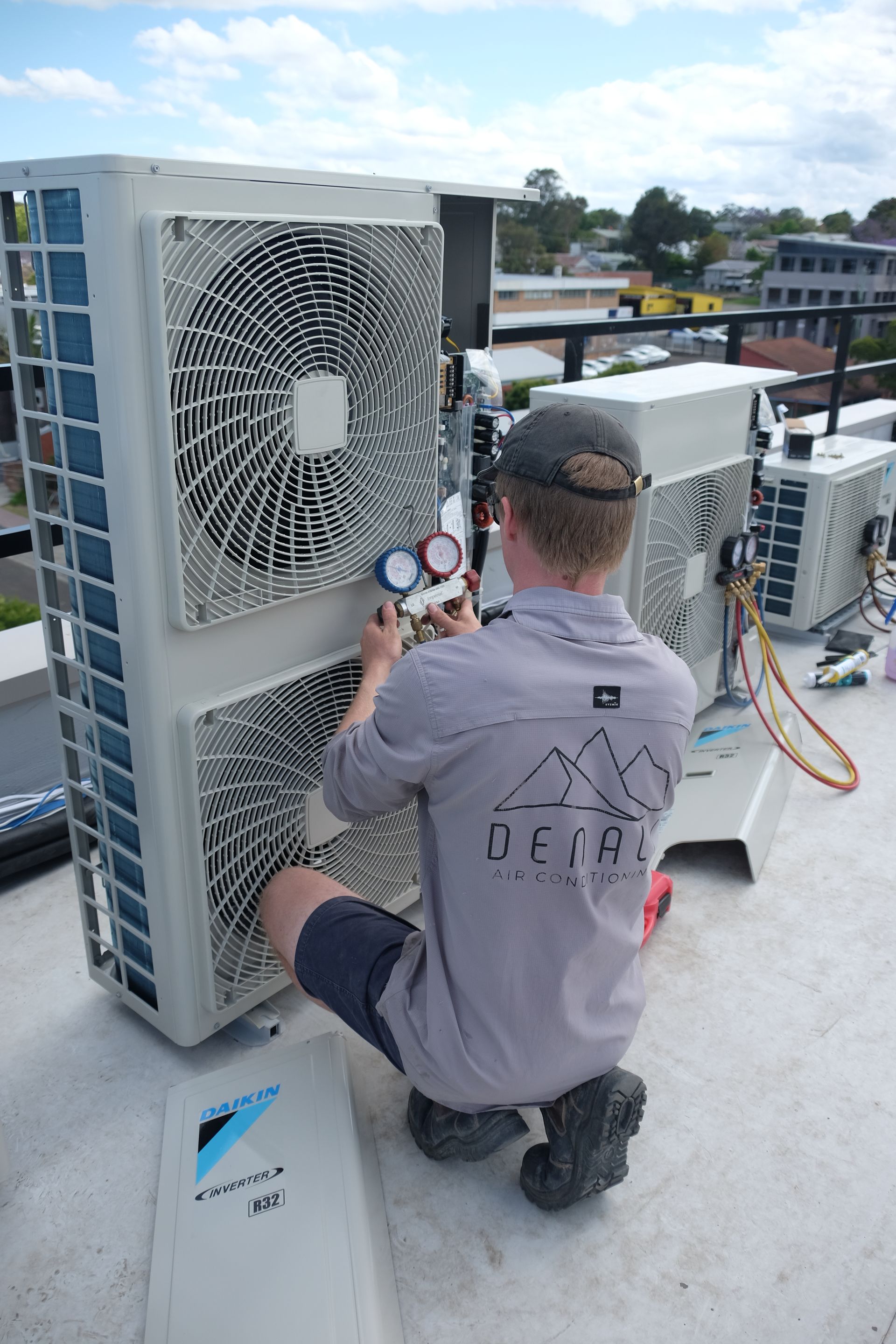 Denali Air Conditioning technician kneels on a rooftop, inspecting air conditioning unit with gauges.
