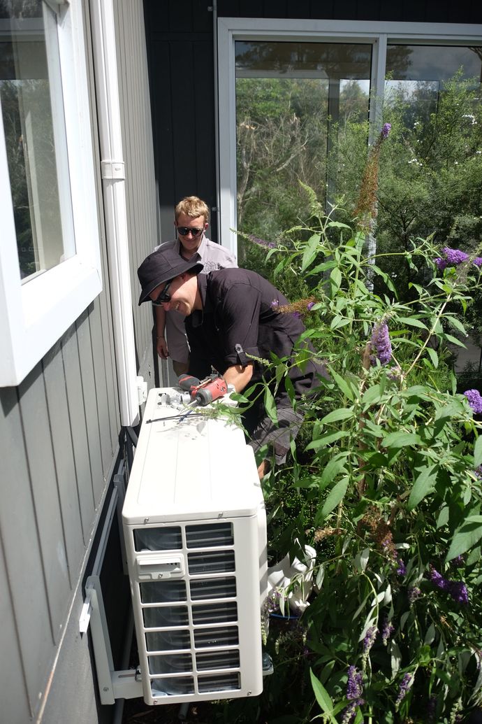 Two Denali Air Conditioning workers in work shirts installing an air conditioner unit next to a building in Nowra