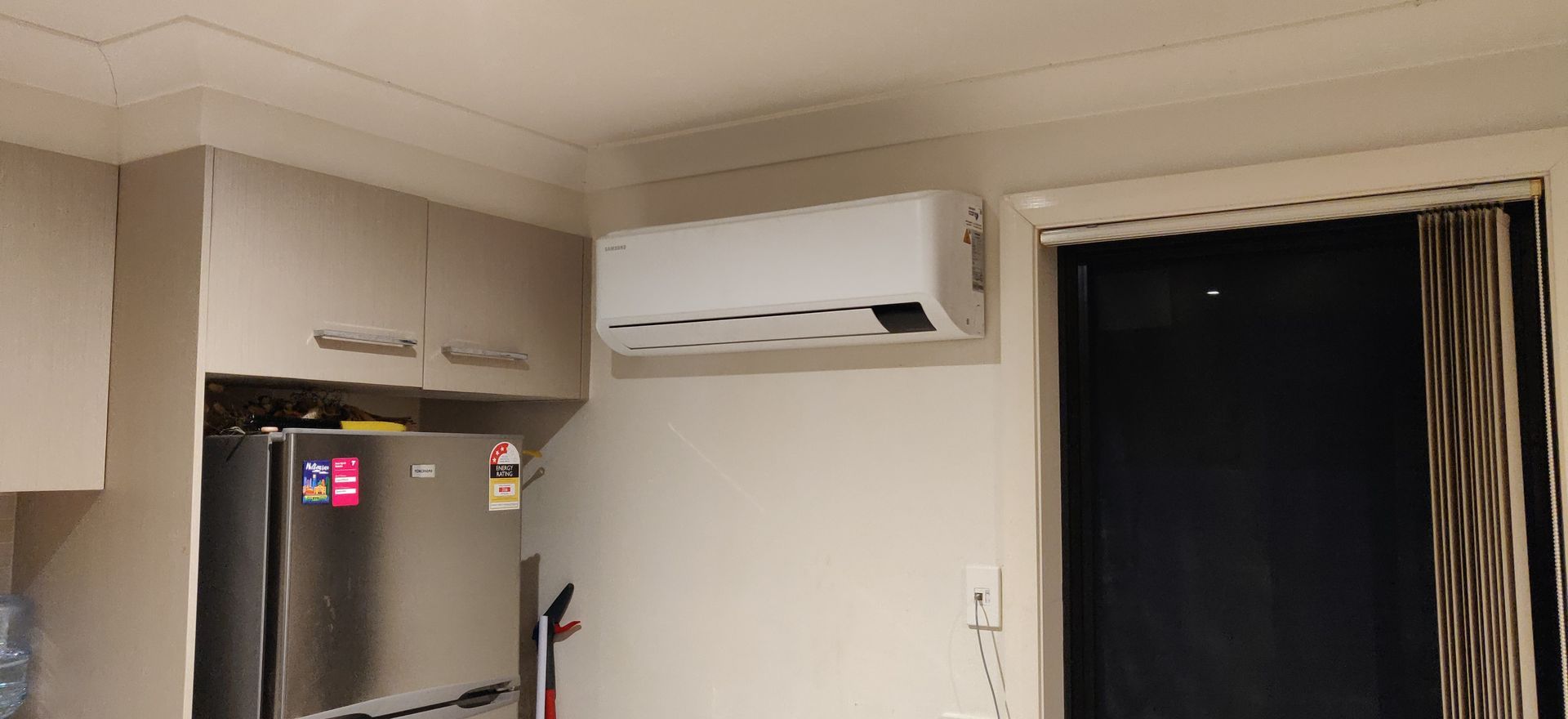 A kitchen with a refrigerator, cabinets, and an air conditioning unit on the wall in Nowra. A window is to the right.