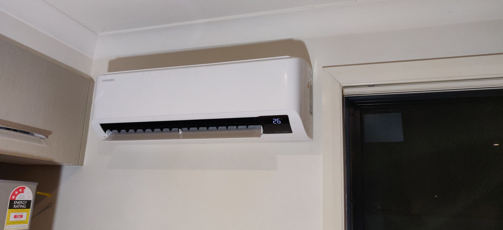 A kitchen with a refrigerator, cabinets, and an air conditioning unit on the wall in Nowra. A window is to the right.