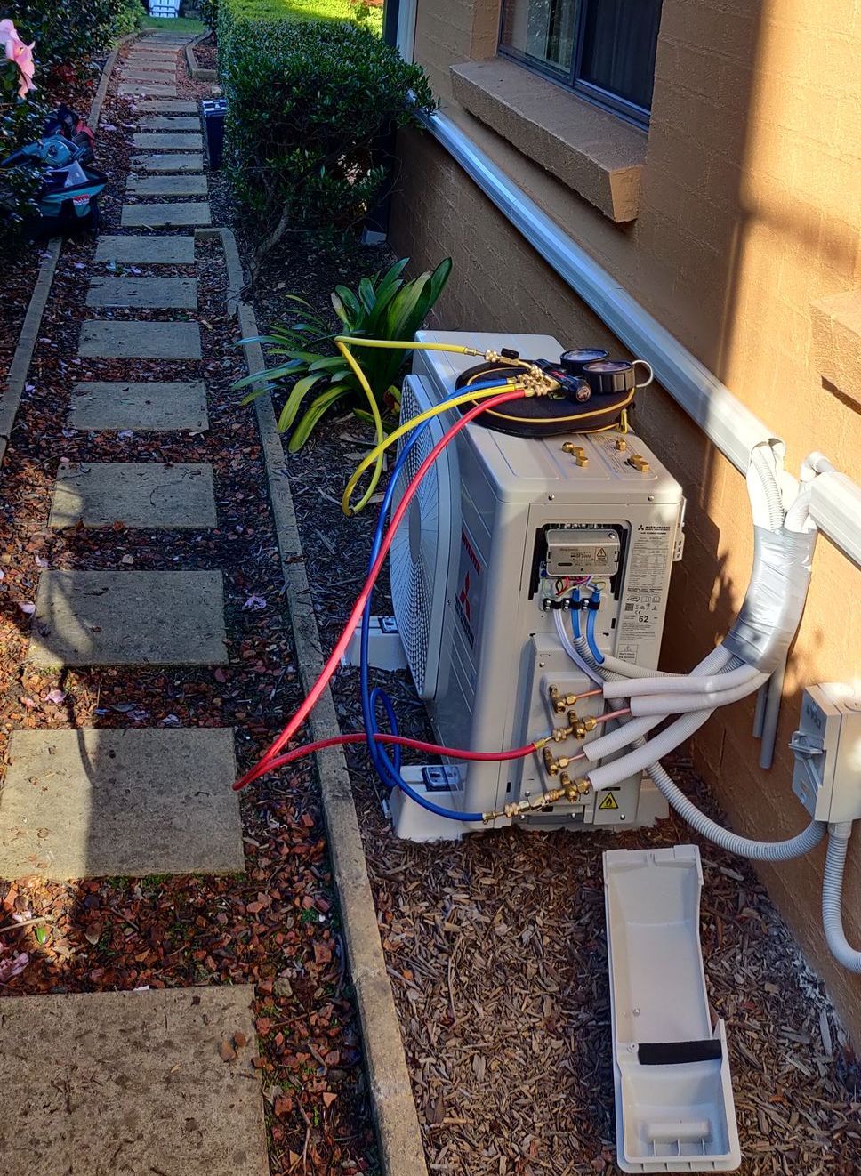 Outdoor AC unit with colored hoses, beside a building and a stepping stone path in Nowra