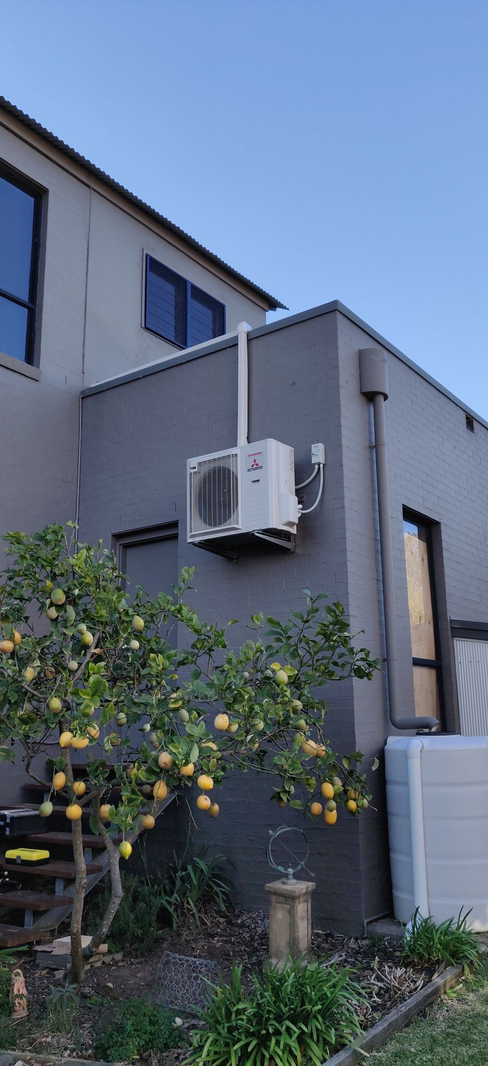 Air conditioning unit in Nowra mounted on a gray building next to a lemon tree under a clear blue sky.