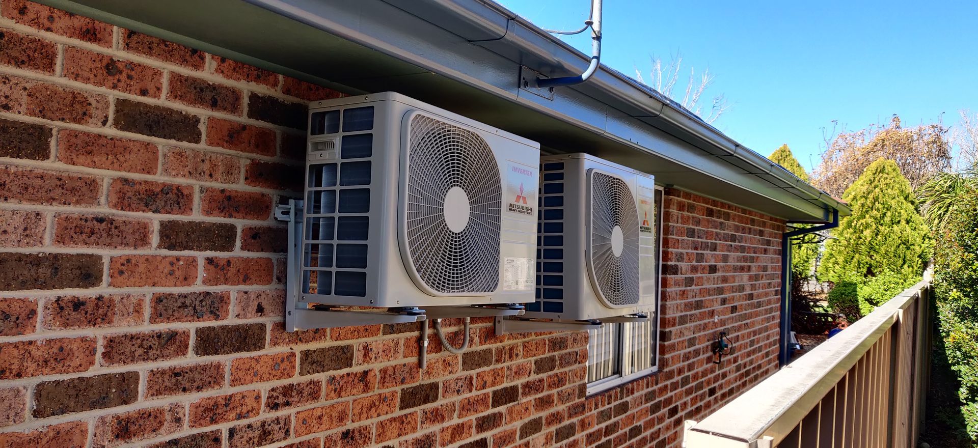 Two air conditioning units mounted on a brick wall under a roof, with a fence and foliage visible in Nowra.