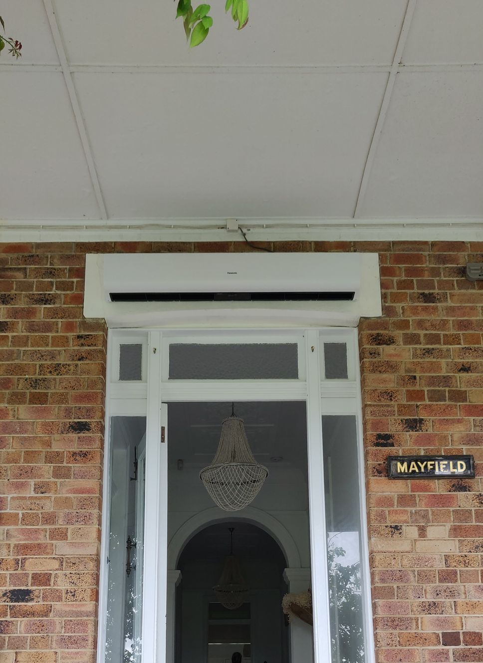 White air conditioner above a white door, brick exterior in Nowra. A chandelier is visible inside.
