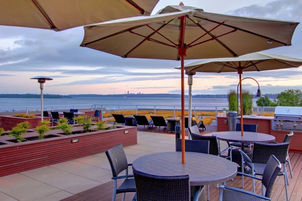 Rooftop patio with umbrellas, tables, and a grill overlooking a bay at dusk.