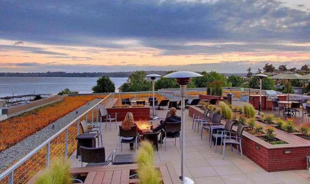 Rooftop patio overlooking a body of water at dusk. People sit near a fire pit, with seating and heaters.