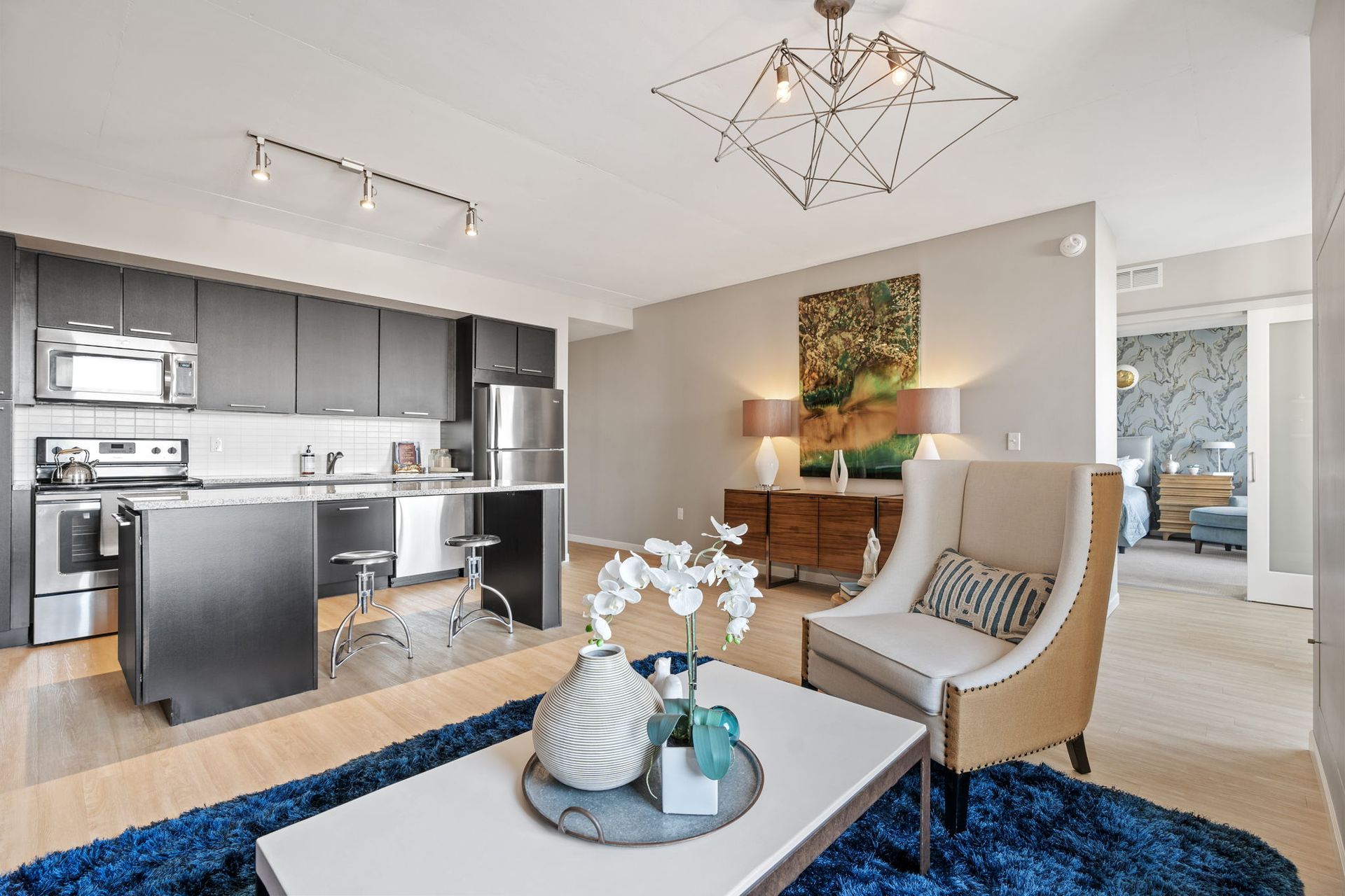 Modern apartment interior with kitchen, living area, and bedroom visible. Blue rug, neutral walls, and contemporary furniture.