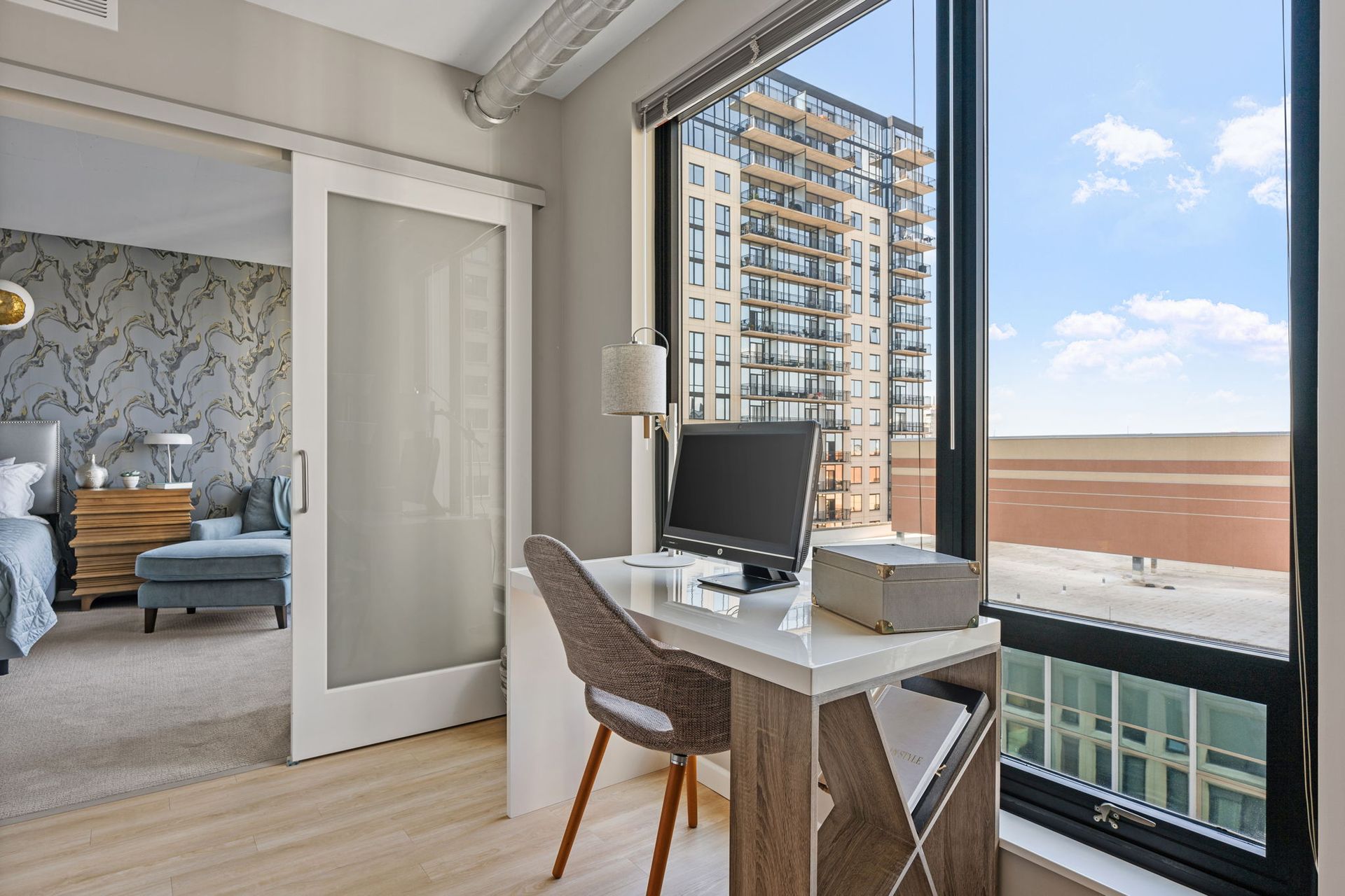Office space with a desk and computer next to a large window overlooking a city. A bedroom is visible through an open door.