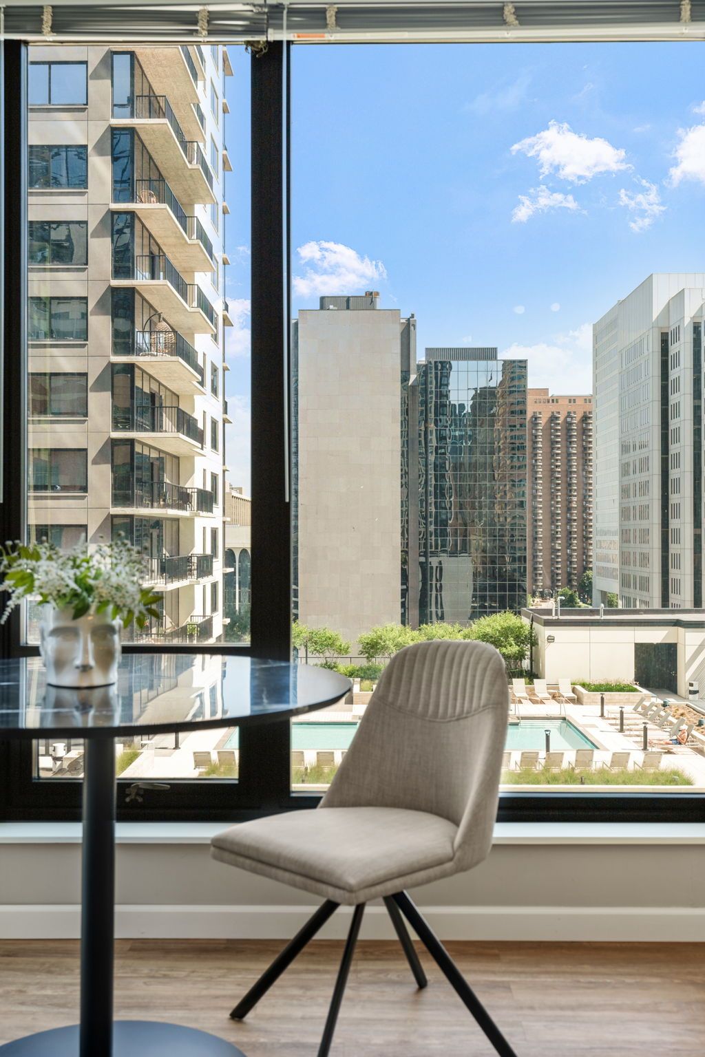A modern dining area with city view. A small table and chair sit near a window, with tall buildings visible outside.