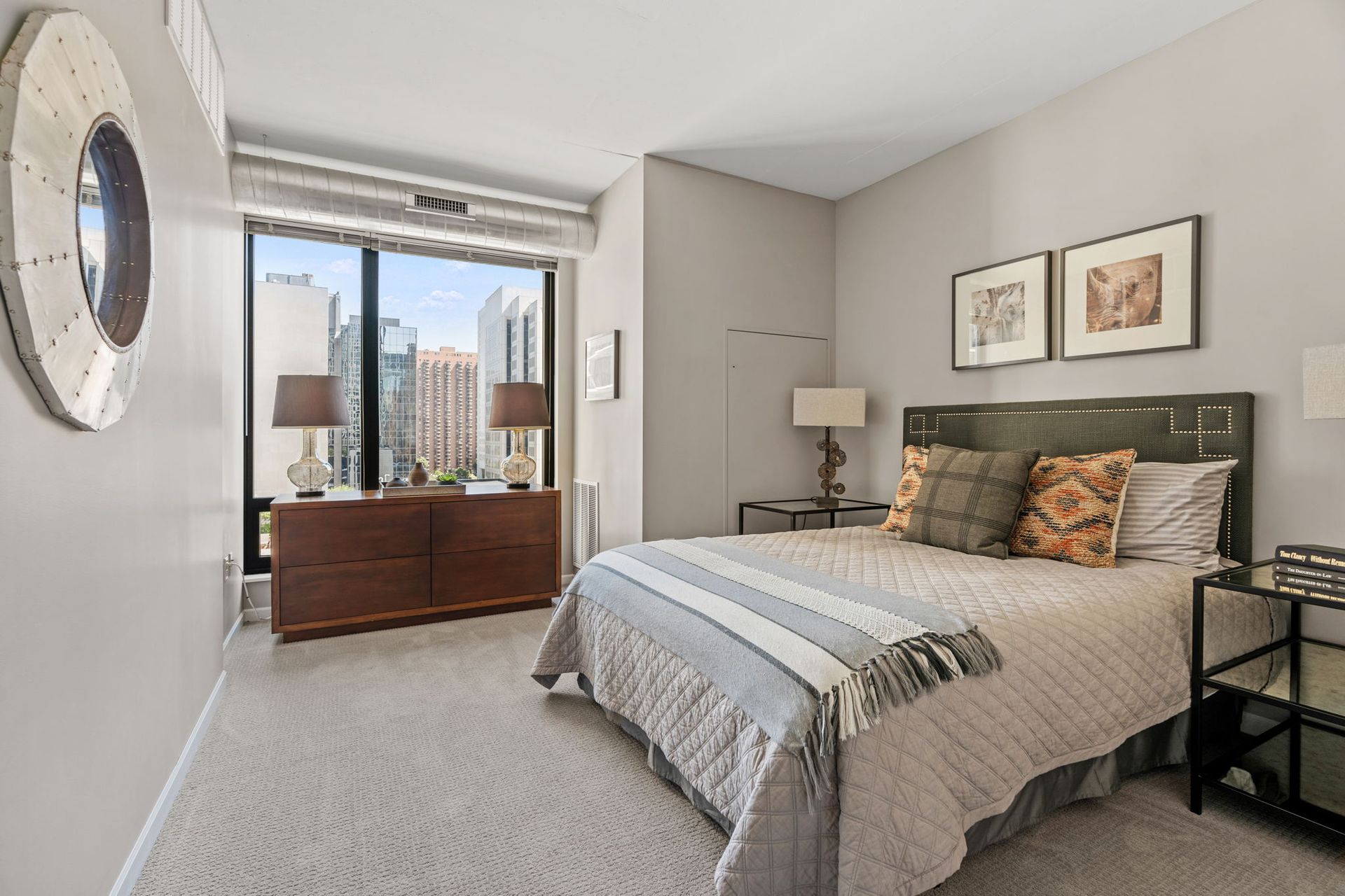 Bedroom with a bed, dresser, and large window overlooking a city. Soft gray and brown color scheme.