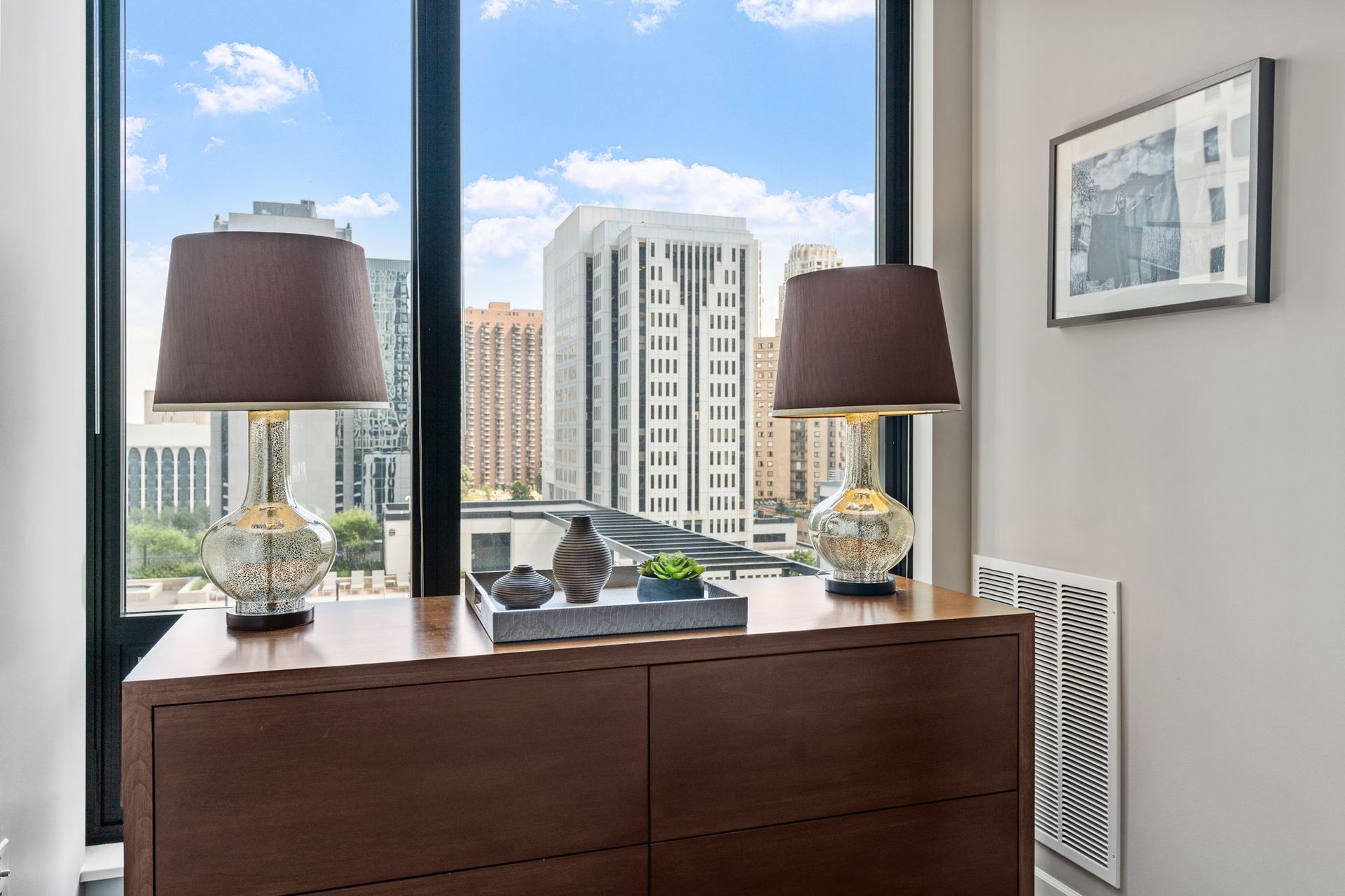 A bedroom with a city view, lamps on a dresser, and artwork on the wall.