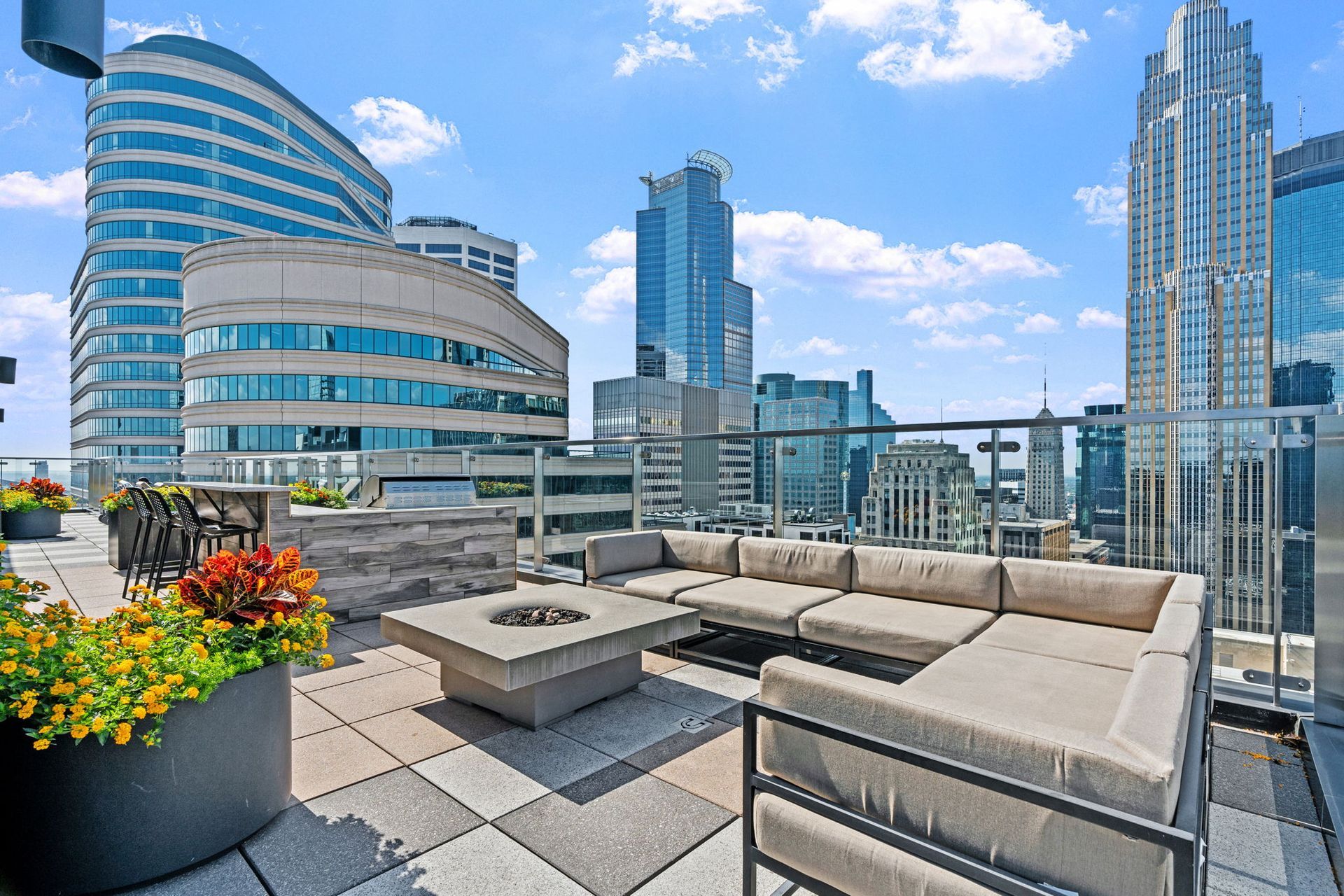 Rooftop patio with modern furniture, a fire pit, and city skyline views on a sunny day.