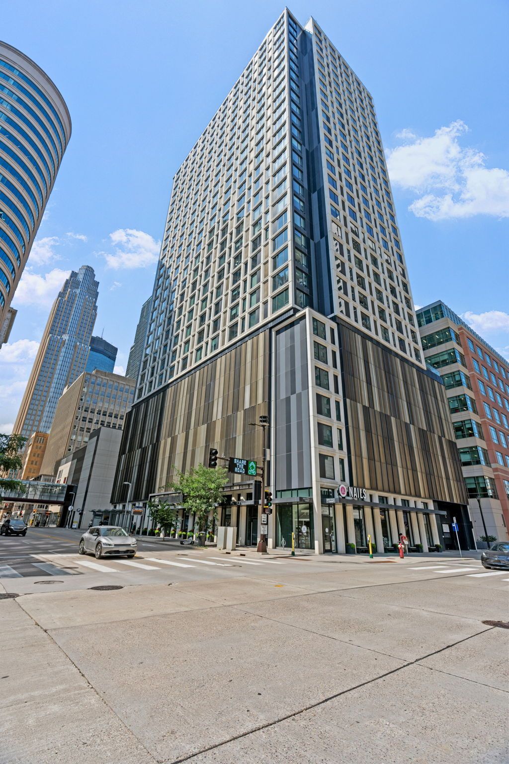 Tall modern building on a city street corner with cars. Blue sky, other buildings visible.